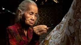 Seorang ibu penjual makanan tradisional tersenyum optimis di pasar, merefleksikan semangat ketahanan ekonomi masyarakat Indonesia. Senyumnya menggambarkan harapan dan upaya gigih dalam merajut kehidupan yang lebih baik.
