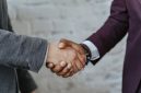 Dua orang berjabat tangan di atas meja yang penuh dengan dokumen, melambangkan kesepakatan bisnis yang kuat dan kolaborasi yang menguntungkan melalui sistem bagi hasil. Ini menunjukkan komitmen bersama untuk mencapai kesuksesan finansial yang adil dan transparan bagi semua pihak terlibat.