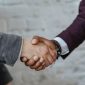 Dua orang berjabat tangan di atas meja yang penuh dengan dokumen, melambangkan kesepakatan bisnis yang kuat dan kolaborasi yang menguntungkan melalui sistem bagi hasil. Ini menunjukkan komitmen bersama untuk mencapai kesuksesan finansial yang adil dan transparan bagi semua pihak terlibat.