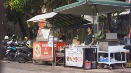 Seorang ibu rumah tangga sedang memilih sayuran di pasar tradisional Indonesia, mencerminkan aktivitas ekonomi sehari-hari serta tantangan dalam memenuhi kebutuhan pokok keluarga. Pasar seperti ini menjadi jantung perekonomian lokal, tempat transaksi dan interaksi sosial yang dinamis terjadi.
